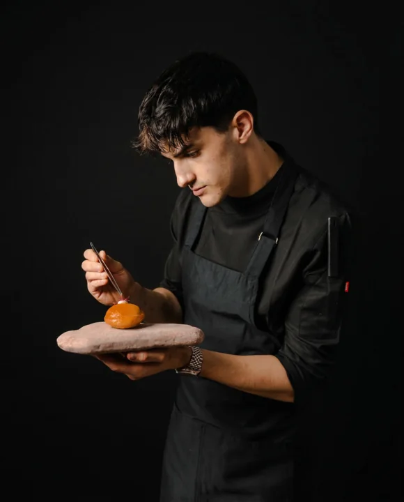 Chef Guillem Roig sosteniendo y terminando un plato sobre fondo oscuro